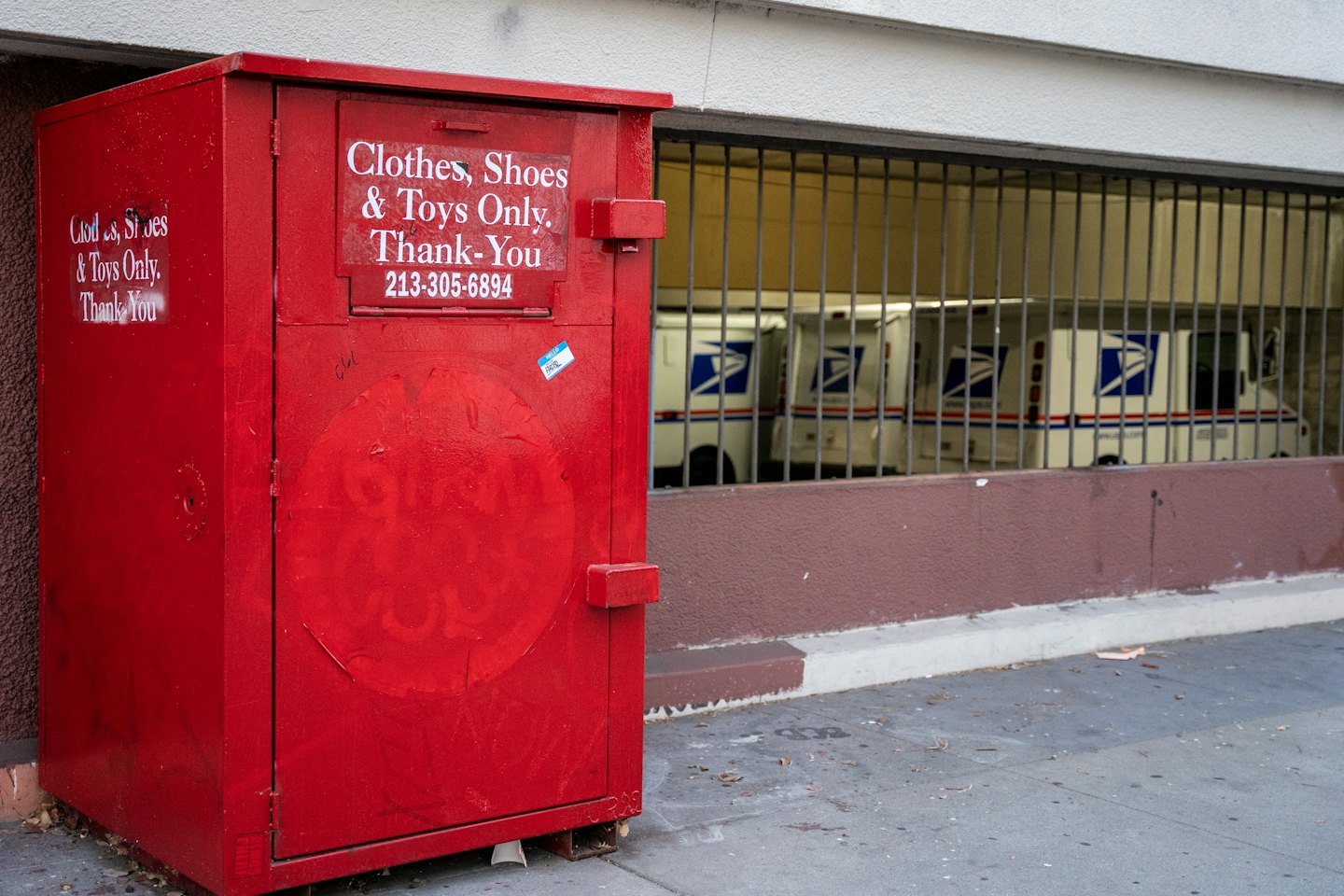 Röd klädinsamlingscontainer på en trottoar med USPS-bilar i bakgrunden.